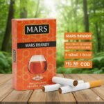 บุหรี่ Mars Brandy ซองสีส้มลายกราฟิกแก้วบรั่นดี พร้อมมวนบุหรี่เม็ดบีบวางบนโต๊ะไม้ฉากหลังป่าสีเขียว
