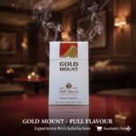 ซองบุหรี่ Gold Mount Red แบบซองแข็ง วางบนโต๊ะไม้สีเข้มในบรรยากาศเลาจน์สุดหรู มีควันลอยละล่องสื่อถึงความเข้มข้นของรสชาติ