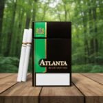 ซองบุหรี่ Atlanta Black Menthol สีดำตัดเขียว วางคู่กับมวนบุหรี่บนโต๊ะไม้ ท่ามกลางบรรยากาศป่าธรรมชาติสีเขียว