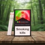 ซองบุหรี่ Marlboro Summer สีดำพร้อมลวดลายสีแดงเขียวและมวนบุหรี่ วางบนโต๊ะไม้ฉากหลังป่า