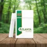 บุหรี่ Atlanta Green Menthol ซองสีขาวแถบเขียวดีไซน์คลาสสิก วางคู่กับมวนบุหรี่บนโต๊ะไม้ ฉากหลังป่าไม้สีเขียว