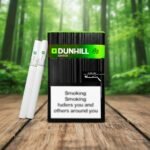บุหรี่ Dunhill Switch Black ซองสีดำแถบเขียว วางคู่กับมวนบุหรี่ก้นกรองชาโคลบนพื้นไม้ ฉากหลังเป็นป่าไม้