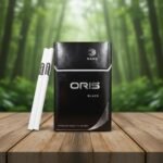 บุหรี่ Oris Nano Black ซองสีดำดีไซน์เรียบหรู วางคู่กับมวนบุหรี่นาโนสลิมบนโต๊ะไม้ ฉากหลังเป็นป่าไม้